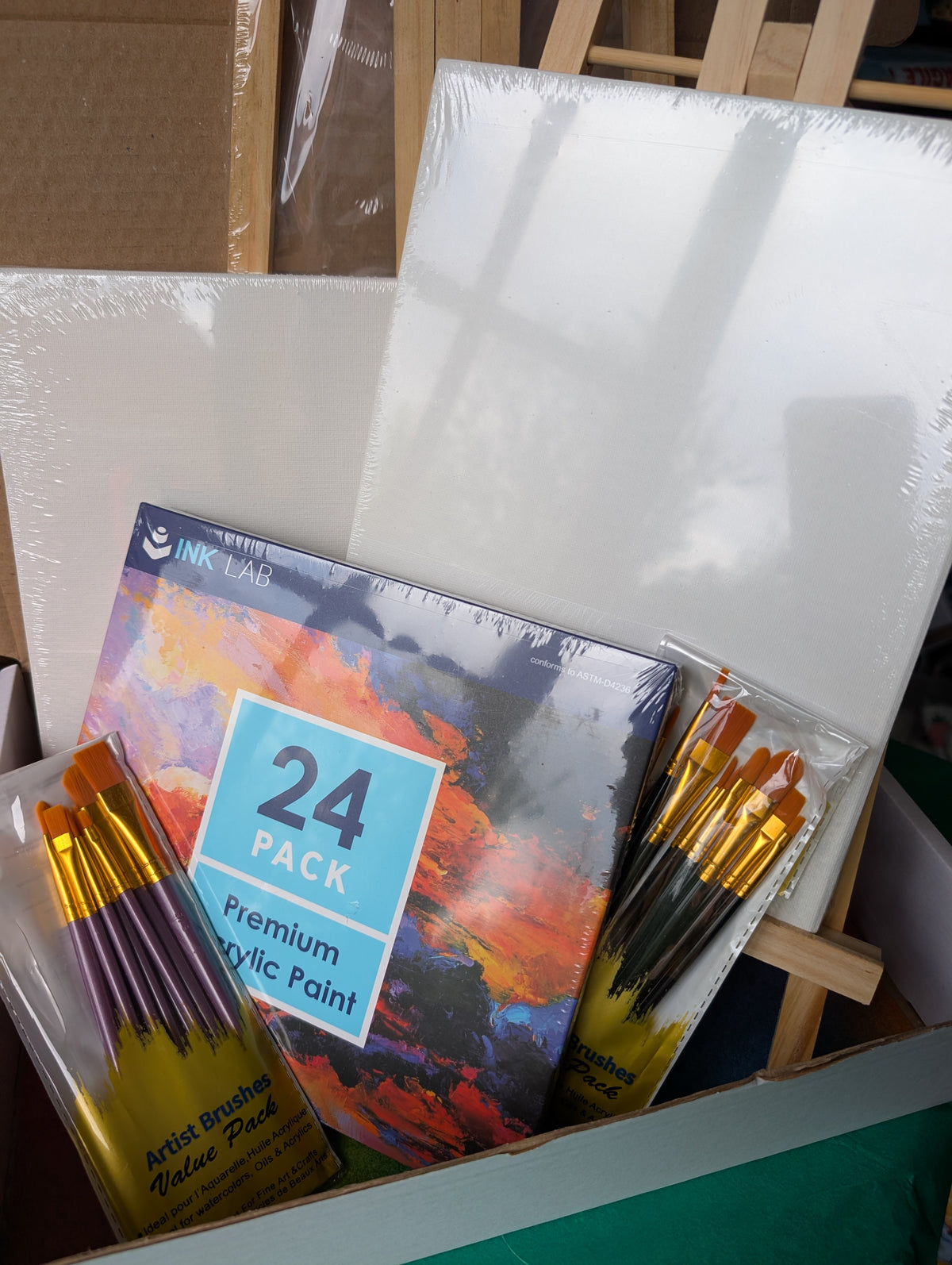 An Ink Lab 24-pack of premium acrylic paint box shown inside a cardboard shipping box alongside two packages of artist brushes and two white canvases on an easel.